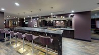 This stylish single-floor amenity space is thoughtfully designed for entertainment and social gatherings, combining multiple functional zones within a cohesive, upscale aesthetic. Rich dark plum and espresso tones dominate the walls and millwork throughout, complemented by wood-look tile flooring and recessed track lighting that creates a warm, lounge-like atmosphere.

At the heart of the space is a striking wet bar featuring an expansive black marble countertop with bold white veining, paired with patterned upholstered bar stools. A large mirrored back bar with dark cabinetry and pendant lighting enhances both the functionality and visual depth of this area.

Adjacent to the bar, a generously sized lounge area offers plush sectional seating arranged around a large flat-screen TV mounted on a textured gold tile accent wall. This zone is bathed in natural light from floor-to-ceiling sliding glass doors that open to an outdoor patio, offering views of the surrounding cityscape and draped with elegant sheer curtains.

The games area includes a full-sized billiards table with a bold red felt surface, set beneath decorative mirrored disco balls that add a playful yet sophisticated flair. A built-in banquette with richly colored cushions provides additional seating nearby. Multi-level step transitions connect the zones, adding architectural dimension to the overall layout.