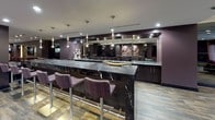 This stylish single-floor amenity space is thoughtfully designed for entertainment and social gatherings, combining multiple functional zones within a cohesive, upscale aesthetic. Rich dark plum and espresso tones dominate the walls and millwork throughout, complemented by wood-look tile flooring and recessed track lighting that creates a warm, lounge-like atmosphere.

At the heart of the space is a striking wet bar featuring an expansive black marble countertop with bold white veining, paired with patterned upholstered bar stools. A large mirrored back bar with dark cabinetry and pendant lighting enhances both the functionality and visual depth of this area.

Adjacent to the bar, a generously sized lounge area offers plush sectional seating arranged around a large flat-screen TV mounted on a textured gold tile accent wall. This zone is bathed in natural light from floor-to-ceiling sliding glass doors that open to an outdoor patio, offering views of the surrounding cityscape and draped with elegant sheer curtains.

The games area includes a full-sized billiards table with a bold red felt surface, set beneath decorative mirrored disco balls that add a playful yet sophisticated flair. A built-in banquette with richly colored cushions provides additional seating nearby. Multi-level step transitions connect the zones, adding architectural dimension to the overall layout.