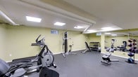 The Grandview Gardens fitness center is a dedicated, single-floor exercise facility designed to support a well-rounded workout routine. The gym features rubber flooring throughout, providing a durable and safe surface ideal for a variety of exercises. The space is equipped with a selection of cardio machines, including a stationary bike and an elliptical trainer, offering residents effective options for cardiovascular workouts. A multi-function cable weight machine serves as a centerpiece for strength training, complemented by an adjustable weight bench and a well-organized dumbbell rack stocked with a range of free weights. A treadmill is also available, rounding out the cardio offerings. The ceiling incorporates exposed ductwork alongside recessed panel lighting, ensuring the space is well-lit throughout. Small ground-level windows allow for modest natural light, adding to the functionality of the environment. The walls are finished in a light neutral green tone, contributing to a clean and energizing atmosphere. Overall, this compact yet practical fitness center provides residents of Grandview Gardens with convenient access to essential exercise equipment within a comfortable and well-maintained setting.