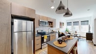 This thoughtfully designed 2-bedroom, 1-bathroom unit at 2939 Main St in Buffalo, NY offers a modern aesthetic with warm, natural wood tones and contemporary finishes throughout. The open-concept kitchen and living area forms the heart of the space, where flat-front wood cabinetry, dark stone countertops, white subway tile backsplash, and a full suite of stainless steel appliances — including a refrigerator, electric range, and microwave — create a functional yet stylish culinary space. A kitchen island with pendant lighting provides additional prep and dining space. Conveniently integrated within the cabinetry is a stacked washer and dryer unit. The living area receives ample natural light through a bank of three windows overlooking the surrounding neighborhood, complemented by warm hardwood flooring that extends throughout. The primary bedroom is a calm, carpeted retreat with a window and dedicated closet space with built-in shelving. The bathroom features warm wood vanity cabinetry with dark stone countertops and hardwood flooring, maintaining a cohesive design language with the rest of the unit. Clean white walls and recessed lighting throughout contribute to a bright, modern ambiance.
