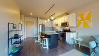 This single-floor residence features a thoughtfully designed open-concept layout that connects the kitchen and living areas seamlessly. The kitchen is a standout space, equipped with stainless steel appliances, including a refrigerator, range, and microwave. White shaker-style cabinetry with matte black hardware pairs with granite countertops, and a penny-tile backsplash adds a distinctive design detail. A dark-toned center island with a granite surface provides bar seating and additional prep space, illuminated by pendant lights and a gold-finish track lighting fixture overhead. Dark hardwood-style flooring runs throughout the main living area.

The living room receives abundant natural light through a large sliding glass door that opens to a private balcony, offering outdoor views of the surrounding landscape. Two bedrooms are carpeted and well-lit by large windows that frame outdoor greenery and open sky views. The bathroom features a granite-topped vanity with white cabinetry, hardwood-style flooring, and a shower-tub combination. Throughout the unit, neutral wall tones, consistent finishes, and modern fixtures contribute to a clean, cohesive aesthetic.