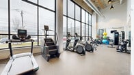 Located at 2171 E 103rd St in Cleveland, Ohio, this fitness center offers a well-appointed, single-floor workout facility designed to accommodate a wide range of exercise needs. The space is defined by its striking floor-to-ceiling windows that line the perimeter, flooding the interior with natural light and providing open views of the surrounding Cleveland cityscape. Soaring ceilings with exposed ductwork and industrial pendant lighting give the space a modern, urban aesthetic, while concrete columns add to its architectural character.

The durable, speckled rubber flooring throughout provides a practical and clean surface suited for high-traffic fitness use. Cardio equipment is prominently featured and includes treadmills, elliptical machines, stair climbers, and stationary bikes, all positioned to take advantage of the expansive window views. A dedicated strength and free weights area offers a dumbbell rack, cable machines, and a bench, complemented by full-length mirrors that enhance the sense of space. Functional training accessories such as medicine balls and stability balls are neatly organized on a storage rack.

Additional amenities include wall-mounted televisions, a built-in water fountain and bottle-filling station, and a private restroom. A glass-paneled entry door provides a welcoming transition into the space. This fitness center is thoughtfully designed to deliver a comprehensive and energizing workout environment.