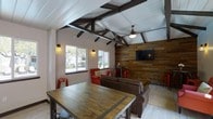 This inviting single-story clubhouse blends rustic charm with modern comfort through a thoughtfully designed interior. The vaulted ceiling immediately draws the eye upward, showcasing dramatic dark wood beam trusses set against a crisp white paneled ceiling, adding architectural character and a sense of openness to the space. A striking reclaimed wood accent wall serves as a natural focal point, warmly textured and earthy in tone, and is outfitted with a mounted flat-screen television for entertainment. The main lounge area is furnished with plush, dark leather seating and vibrant red accent chairs and stools, creating a bold, welcoming color contrast. A large communal wood table with metal accents anchors the room, making it ideal for gatherings, meetings, or casual socializing. Wall-mounted light sconces and a ceiling fan with a pendant light provide both functional illumination and a cozy ambiance throughout. Natural light flows in generously through a set of large windows that offer views of the surrounding outdoor greenery, further enhancing the warm and relaxed atmosphere. The overall design strikes a balance between rustic aesthetics and contemporary amenities, making this clubhouse a comfortable and functional shared space.