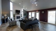 This single-floor space makes a strong visual impression with its vaulted ceilings and thoughtfully curated interior design. The main living area serves as the heart of the space, anchored by a striking white ornate fireplace with a mounted television above — a bold focal point framed by white-painted brick and flanked by decorative topiary accents. The ceiling rises to a generous peak, enhancing the open and airy feel of the room, while a ceiling fan ensures comfort throughout. Deep plum-colored accent walls contrast elegantly against the gray and white tones found elsewhere, lending the space a rich and sophisticated ambiance. Stone-look tile flooring runs throughout, providing a durable yet attractive foundation that complements the overall aesthetic. Large windows and glass doors along one wall flood the interior with natural light, creating a bright and inviting atmosphere. The living area is furnished with dark leather seating, patterned armchairs, and a classic area rug, offering a balanced mix of comfort and style. A large wall clock adds a decorative touch to the accent wall, completing the well-appointed look of this distinctive and functional space.