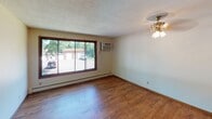 This single-floor space offers a functional and straightforward layout, highlighted by warm hardwood-style flooring that runs consistently throughout the main living areas. The living room is a generous, open area bathed in natural light from a large sliding glass window that frames outdoor views of surrounding greenery. A ceiling fan with a light fixture provides both comfort and illumination, while a wall-mounted air conditioning unit and baseboard heating ensure year-round climate control. Textured ceilings are present throughout the unit.

The kitchen features a peninsula layout with a light-colored laminate countertop and rich, dark wood cabinetry extending to the ceiling, offering ample storage. Essential appliances include a refrigerator and an electric range. The peninsula serves as a natural divider between the kitchen and the living space, creating an open, connected feel.

The bedroom is a quiet, well-proportioned room with neutral cream-toned walls and the same hardwood-style flooring, providing a clean and versatile space. The bathroom includes white square tile wainscoting, a tub-shower combination with a curtain, a toilet, and a wood-framed vanity with a large mirror and overhead lighting, offering a practical and tidy design.
