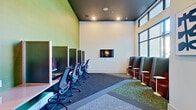 This single-floor business center is thoughtfully designed to accommodate a variety of work and collaboration needs. The space is divided into two distinct functional zones that complement each other seamlessly. Along one wall, a row of individual computer workstations is set against a bold green accent wall, each station separated by privacy panels and outfitted with ergonomic mesh chairs, creating a focused environment for productive work. Opposite this area, a series of high-backed leather lounge pods — upholstered in rich burgundy and charcoal — are arranged along expansive floor-to-ceiling windows, each accompanied by a small white side table, offering a more relaxed yet private setting for individual work or casual meetings. The oversized windows flood the space with natural light and provide views of the surrounding urban streetscape. A wall-mounted flat-screen display serves as a central focal point, adding a media-ready element to the room. The dark ceiling with recessed lighting creates a sleek contrast against the white and green walls, while patterned carpet tiles in varying textures define each seating zone. Curated artwork adorns the walls near the entry, adding a refined, creative touch. Glass-paneled doors connect the space to adjacent areas, enhancing its open, connected feel.