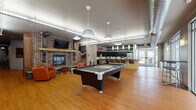 Located on S Broadway Street in Akron, Ohio, this single-floor communal space blends industrial aesthetics with comfortable, functional design. The open layout is unified by warm hardwood flooring that runs throughout, creating a cohesive flow between distinct activity zones.

The lounge area anchors one end of the space, centered around a striking exposed brick fireplace wall with a mounted flat-screen television and orange upholstered seating. Nearby, a full-size pool table serves as a focal point for recreation. Pendant lighting with industrial-style fixtures hangs from the ceiling, complementing exposed ductwork that reinforces the building's urban character.

A casual seating nook features patterned armchairs arranged around a low table, positioned near a staircase with wood and metal railings. Large floor-to-ceiling windows and glass walls flood this area with natural light while offering views of the surrounding exterior.

The kitchen area is well-appointed with dark espresso cabinetry, granite countertops, stainless steel appliances including a refrigerator and dishwasher, and an island with bar seating. A bold "EAT" sign adds a playful touch to the space. The combination of brick, exposed ducts, and modern finishes gives the entire space a polished yet welcoming industrial character.