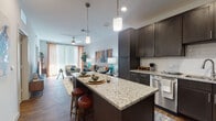 This thoughtfully designed single-floor residence showcases a seamless open-concept layout that connects the kitchen, dining, and living areas into one cohesive, light-filled space. The kitchen is a standout, featuring rich dark espresso cabinetry, granite countertops, a white subway tile backsplash, and a full suite of stainless steel appliances including a refrigerator, range, microwave, and dishwasher. A spacious central island with additional open shelving below provides both extra prep space and casual seating, making it ideal for entertaining. Warm wood-tone flooring runs throughout the main living areas, complementing the kitchen's dark cabinetry and adding depth to the overall aesthetic. The living room is generously proportioned, filled with natural light from large floor-to-ceiling windows dressed with vertical blinds and sheer curtains. A ceiling fan overhead adds comfort, while the open wall space invites personalized décor. The bedroom offers a comfortable and private retreat, carpeted for warmth, with a window allowing in natural light. It connects directly to a well-organized walk-in closet with built-in shelving and hanging space. The bathroom features light-toned tile flooring, a granite-topped dark vanity, and clean, modern fixtures, completing this polished and functional living space.