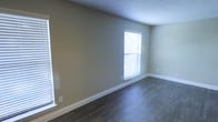 This single-floor space presents a straightforward and functional interior design. The main room visible features dark hardwood-style flooring that provides a rich contrast against the neutral, light sage-green painted walls. White baseboards run along the perimeter of the room, adding a crisp, clean finish to the overall aesthetic.

Two windows fitted with white horizontal blinds allow natural light to filter into the space, brightening the room while offering privacy control. The windows are positioned to provide good light coverage across the room. The walls are smooth and freshly painted, offering a versatile backdrop suitable for a variety of furnishing styles.

The ceiling is standard height, contributing to a comfortable and proportionate feel within the room. The space is unfurnished, allowing for a clear view of the flooring and architectural details. The overall condition of the space appears well-maintained, with clean lines and minimal wear visible on both the floors and walls. The combination of dark flooring, light walls, and white trim creates a balanced, neutral palette that would suit a range of uses and interior design preferences.