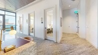 Located in the heart of Nashville on 8th Avenue South, this polished commercial space combines a sophisticated lounge area with functional private workspaces. The open lounge is anchored by a stylish patterned carpet and outfitted with a mix of freestanding tables and white modern chairs, complemented by built-in banquette seating upholstered in warm yellow tones. Overhead, a striking slatted wood ceiling detail adds warmth and architectural character, while elegant pendant lights with white drum shades provide focused illumination. Large floor-to-ceiling windows flood the space with natural light and offer impressive views of the Nashville skyline, creating an energizing and inspiring atmosphere. A service counter with a marble-topped surface adds a refined, hospitality-forward touch. Curated artwork adorns the crisp white walls, lending a creative ambiance throughout. Toward the rear, a corridor leads to several private offices and focus rooms, each accessed through glass-paneled doors framed in white millwork and accented by decorative brass wall sconces. These compact individual offices are equipped with desks and ergonomic seating, ideal for focused work. The overall design balances professional functionality with an elevated, welcoming aesthetic suited for a modern coworking or amenity environment.