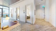 Located in the heart of Nashville on 8th Avenue South, this polished commercial space combines a sophisticated lounge area with functional private workspaces. The open lounge is anchored by a stylish patterned carpet and outfitted with a mix of freestanding tables and white modern chairs, complemented by built-in banquette seating upholstered in warm yellow tones. Overhead, a striking slatted wood ceiling detail adds warmth and architectural character, while elegant pendant lights with white drum shades provide focused illumination. Large floor-to-ceiling windows flood the space with natural light and offer impressive views of the Nashville skyline, creating an energizing and inspiring atmosphere. A service counter with a marble-topped surface adds a refined, hospitality-forward touch. Curated artwork adorns the crisp white walls, lending a creative ambiance throughout. Toward the rear, a corridor leads to several private offices and focus rooms, each accessed through glass-paneled doors framed in white millwork and accented by decorative brass wall sconces. These compact individual offices are equipped with desks and ergonomic seating, ideal for focused work. The overall design balances professional functionality with an elevated, welcoming aesthetic suited for a modern coworking or amenity environment.