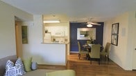 This single-floor residence offers an open-concept design that seamlessly connects the kitchen, dining, and living areas into one cohesive and functional space. The kitchen features white cabinetry, white appliances, and a granite-topped breakfast bar that serves as a natural divider between the cooking and living zones, providing both workspace and casual seating potential. Under-cabinet lighting adds a practical touch to the kitchen area.

The dining area is a standout feature, highlighted by a bold navy accent wall that creates a striking visual contrast against the lighter tones throughout the space. A ceiling fan with a light fixture overhead provides both airflow and ambient lighting. A wall-mounted mirror enhances the sense of depth in the dining area, while a round dining table surrounded by green upholstered chairs adds a contemporary, welcoming feel.

Warm hardwood-style flooring flows throughout the main living areas, adding continuity and warmth to the space. The textured ceiling adds a classic finish, while the neutral wall tones balance the bolder design elements. Together, these details create an inviting, well-appointed interior suited for everyday living and entertaining.