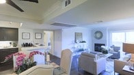 This single-floor residence offers an open-concept layout that effortlessly connects the living, dining, and kitchen areas into one cohesive, light-filled space. The interior is finished with crisp white walls and crown molding that runs throughout, lending the home a refined, classic character. Dark hardwood flooring grounds the space and provides a striking contrast to the bright, airy palette above.

The living area is anchored by a sleek fireplace flanked by decorative accents, creating a warm and inviting focal point. A stylish console table and oversized wall clock add personality and charm to the space. Generous ceiling height, enhanced by a tray ceiling detail, contributes to an expansive sense of volume.

The open kitchen features a clean white design with bar-height seating, making it equally functional for casual dining and entertaining. It flows naturally into the dining area, where upholstered chairs with nailhead trim add a touch of sophistication.

Natural light fills the space through multiple vents and openings, keeping the atmosphere bright throughout the day. A hallway visible from the main living area suggests additional private rooms beyond. Overall, this residence combines modern elegance with comfortable livability in a thoughtfully designed single-level layout.
