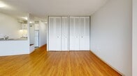 Located on Marine Drive in North Vancouver, BC, this efficiently designed studio unit makes smart use of its single-floor layout. The open-concept main area seamlessly combines the living, dining, and kitchen functions into one cohesive space, creating a practical and flexible environment. Warm hardwood-style flooring runs throughout the living area, complementing the bright, neutral walls and textured ceilings.

Large windows dominate the living area, flooding the space with natural light and offering views of the surrounding North Vancouver cityscape and distant mountain backdrop — a standout feature that adds a sense of openness well beyond the unit's footprint. Baseboard heating runs along the window walls, ensuring year-round comfort.

The kitchen is positioned adjacent to the main living area and features white cabinetry with a refrigerator, providing functional meal preparation space. The full bathroom is well-appointed with a single vanity featuring white cabinetry and a countertop with an integrated sink, a large mirror, and a tub-shower combination with clean white tile surround and tile flooring.

A notable highlight is the extensive closet storage, with multiple sets of louvred bifold closet doors offering an impressive amount of built-in storage space — a practical advantage in a compact unit. The overall ambiance is clean, bright, and move-in ready.