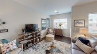 This single-floor residence offers a thoughtfully designed layout with a contemporary aesthetic carried consistently throughout each space. The kitchen serves as a functional centerpiece, featuring flat-front cabinetry in a warm taupe finish, stainless steel appliances including a gas range, built-in microwave, and French door refrigerator. A large island with a white quartz countertop provides bar seating, illuminated by pendant lighting overhead. Dark hardwood-style flooring runs through the kitchen and living areas, grounding the neutral color palette.

The open living room receives generous natural light through multiple windows dressed with horizontal blinds, offering views of the surrounding neighborhood. The space comfortably accommodates a full sectional and additional seating, creating a welcoming, versatile area.

The primary bedroom is notably well-lit, with wide windows along one wall and soft carpet underfoot. A second bedroom mirrors this bright, calm atmosphere. Both rooms feature neutral wall tones and ample space for furnishings.

The bathroom includes large-format tile flooring, a wood-toned vanity with white countertop, a wall-mounted toilet, and a glass-enclosed walk-in shower, delivering a clean, modern finish throughout.