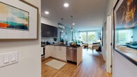 This well-appointed single-floor residence offers a thoughtfully designed open-concept layout that seamlessly connects the kitchen, dining, and living areas. The kitchen is a standout feature, equipped with stainless steel appliances including a French door refrigerator, built-in microwave, and range. Two-tone cabinetry — dark upper cabinets paired with crisp white lower cabinets — is complemented by dark countertops and a tile backsplash. A central island with seating provides additional prep and entertaining space, while elegant pendant lighting adds a modern touch overhead. The open living area is bright and inviting, with large windows that flood the space with natural light and offer views of lush greenery. Light hardwood flooring flows consistently throughout the unit, enhancing the sense of continuity. The bedroom features a bold accent wall in a deep slate blue, creating a cozy and stylish retreat, with a window framing outdoor views. The bathroom includes a soaking tub with full tile surround and a dark-countered vanity. A dedicated in-unit laundry area with a stacked washer and dryer adds everyday convenience. A wall-mounted mini-split system provides climate control throughout.