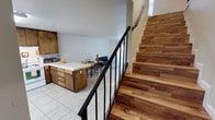 This two-story space is designed for functional, everyday living with a layout that seamlessly connects its main living areas. The ground floor opens into an open-concept kitchen, dining, and living zone, creating a cohesive and welcoming environment. The kitchen features warm-toned wood cabinetry, a tile-top island with built-in storage drawers, and light-colored tile flooring that extends throughout the main level. Adjacent to the kitchen, a dining area with a small table provides a casual space for meals, accented by a cheerful touch of fresh flowers. The living area just beyond offers comfortable seating and a relaxed atmosphere. A striking hardwood staircase with rich, natural wood grain tones and a sleek black metal railing serves as a visual centerpiece, connecting the ground floor to the upper level. The clean white walls throughout keep the space feeling bright and airy, complementing the natural warmth of the wood elements. The upper floor provides additional living or sleeping quarters, accessible via the prominently featured staircase. Overall, the space balances practicality with inviting design details, making it a comfortable and well-organized multi-level home.