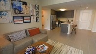 This single-floor space showcases a thoughtfully designed open-concept layout that seamlessly connects the living and kitchen areas, creating a cohesive and functional environment. The living area is anchored by a plush sectional sofa adorned with bold accent pillows in rust and patterned tones, complemented by a rustic wooden coffee table that adds warmth to the space. A multicolored area rug defines the seating zone, while an art-forward gallery wall featuring eclectic framed prints adds personality and visual interest.

The kitchen is a standout feature, boasting dark cabinetry with a sleek, modern aesthetic, stainless steel appliances including a French door refrigerator, and a central island with bar seating. Light countertops contrast beautifully against the dark cabinetry, creating a sophisticated two-tone palette. The flush-mounted ceiling light provides warm, even illumination throughout the cooking space.

Large-format tile flooring runs throughout, lending a clean, contemporary feel and creating visual continuity between spaces. A hallway visible in the background hints at additional rooms beyond, while neutral wall tones keep the overall ambiance bright and inviting.