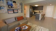 This single-floor space showcases a thoughtfully designed open-concept layout that seamlessly connects the living and kitchen areas, creating a cohesive and functional environment. The living area is anchored by a plush sectional sofa adorned with bold accent pillows in rust and patterned tones, complemented by a rustic wooden coffee table that adds warmth to the space. A multicolored area rug defines the seating zone, while an art-forward gallery wall featuring eclectic framed prints adds personality and visual interest.

The kitchen is a standout feature, boasting dark cabinetry with a sleek, modern aesthetic, stainless steel appliances including a French door refrigerator, and a central island with bar seating. Light countertops contrast beautifully against the dark cabinetry, creating a sophisticated two-tone palette. The flush-mounted ceiling light provides warm, even illumination throughout the cooking space.

Large-format tile flooring runs throughout, lending a clean, contemporary feel and creating visual continuity between spaces. A hallway visible in the background hints at additional rooms beyond, while neutral wall tones keep the overall ambiance bright and inviting.