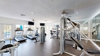 This fitness center is a comprehensively outfitted single-level space designed to support a full range of exercise needs. The layout is thoughtfully organized, with cardio equipment — including multiple treadmills and elliptical machines — positioned along the windowed perimeter, allowing users to enjoy natural light and views of the adjacent outdoor pool area while working out. French doors provide additional access points and further enhance the brightness of the space.

The strength training section is well-appointed with Inflight Fitness cable machines and multi-function weight stations, while a dedicated free weight area includes a dumbbell rack for supplemental training. Ceiling fans support ventilation and comfort throughout the space, complemented by a standard HVAC system. Multiple wall-mounted flat-screen televisions are positioned for easy viewing from various workout stations, adding an entertainment element to the fitness experience.

Full-length mirrors line one wall, visually expanding the space and providing users with form-monitoring capabilities. The durable rubber flooring is practical and appropriate for a high-use fitness environment. A small side room with a service window and fire safety equipment adds functional utility. The overall design is clean and functional, with white walls and ample lighting contributing to an open, energizing atmosphere.
