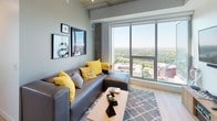 This contemporary single-floor residence blends industrial and modern design elements throughout its well-appointed layout. Exposed concrete ceilings and structural columns contrast beautifully with light hardwood-style flooring, creating a refined urban aesthetic that flows through the common areas.

The galley-style kitchen is thoughtfully designed with white upper cabinetry, dark lower cabinets, and a striking chevron-patterned tile backsplash. Stainless steel appliances — including a refrigerator, electric range, built-in microwave, and dishwasher — provide full functionality, while a breakfast bar with seating adds a casual dining option.

The living area is bright and airy, anchored by a large floor-to-ceiling window that frames sweeping panoramic views of the city skyline and lush treetops below, flooding the room with natural light.

The two bedrooms feature carpeted flooring, generous closet storage with open shelving and hanging rails, and windows offering the same impressive elevated views. Each room accommodates a comfortable workspace alongside sleeping quarters.

Two well-appointed bathrooms serve the residence. One features a soaking tub with tiled surround, while the other offers a glass-enclosed walk-in shower. Both include dark wood vanities, white quartz countertops, and large mirrors.