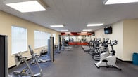 This single-floor fitness center offers a spacious and functional layout designed to accommodate a wide range of workout needs. The space features a durable rubberized flooring throughout, providing a safe and practical surface for exercise. Overhead, a drop tile ceiling with recessed fluorescent panel lighting ensures the area is brightly and evenly illuminated.

The cardio section is well-stocked with multiple treadmills, ellipticals, and stationary bikes, arranged in rows to maximize floor space and usability. A mounted flat-screen television is positioned on the wall, offering entertainment during workouts. The strength training area includes a variety of cable machines and weight equipment, catering to users at different fitness levels.

A bold red accent wall with a large mirror spans one end of the room, visually expanding the space while allowing users to monitor their form. Natural light filters in through windows fitted with white blinds along the side wall, complementing the artificial lighting. A wall-mounted HVAC unit ensures climate control for year-round comfort, making this fitness center a practical and inviting environment for exercise.