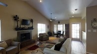 This single-floor home offers a welcoming and well-designed open-concept layout that seamlessly connects the living and dining areas. The living room is a standout space, highlighted by soaring vaulted ceilings that create an impressive sense of height and openness. A tiled fireplace with a dark mantel serves as a natural focal point, complemented by a ceiling fan that adds both function and style. Recessed lighting throughout ensures a well-lit interior, while multiple large windows dressed with white blinds invite generous amounts of natural light. A glass-paneled door provides easy access to the outdoors and further brightens the space. The hardwood floors extend throughout, adding warmth and continuity. The adjacent dining area accommodates a table and chairs and benefits from the same open, airy atmosphere. The neutral wall tones and warm finishes throughout give the home a cohesive, inviting ambiance. Overall, this home combines practical design with comfortable living spaces, making it a versatile and charming single-level residence.