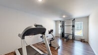 This single-floor amenity space is thoughtfully organized into distinct functional areas, offering both fitness and laundry facilities in a clean, well-maintained environment. The fitness room serves as the centerpiece, featuring hardwood-style flooring, recessed lighting, and crisp white walls that create a bright and motivating atmosphere. The room is equipped with a Precor treadmill, an upright stationary bike, and a multi-station cable strength machine, providing a well-rounded selection of cardio and strength training options. A window along one wall allows natural light to filter in, adding to the open feel of the space. An adjacent area provides additional open floor space, suitable for stretching or floor exercises. The laundry room is a practical and efficiently arranged space, housing multiple front-loading washer and dryer units along the walls, making it capable of handling high-volume use. It shares the same durable hardwood-style flooring and neutral wall finishes seen throughout, maintaining a consistent and tidy aesthetic. A fire extinguisher is visibly mounted in the laundry room, reflecting attention to safety. The layout flows logically between areas via a connecting hallway, and the overall design prioritizes functionality, cleanliness, and ease of use.