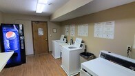 This shared laundry facility is a single-floor space designed with practicality and convenience in mind. The room features a row of white top-load washing machines and dryers arranged along the walls, providing ample capacity for multiple users. The warm-toned beige walls are lined with informational posters offering laundry tips, usage instructions, and service contact details, ensuring residents have all the guidance they need at a glance. The space is finished with durable wood-look flooring that is easy to clean and maintain, complemented by a textured ceiling with overhead fluorescent lighting that keeps the room bright and functional. A wooden entry door with posted notices provides a clear access point to the facility. A trash receptacle is conveniently placed for easy disposal of lint and packaging materials. The overall layout is straightforward and efficient, maximizing usable floor space while maintaining a tidy, organized environment. The room's neutral color palette and clean design contribute to a no-fuss, utilitarian ambiance suited for its purpose as a communal laundry space.