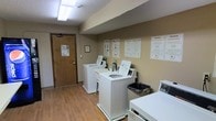This shared laundry facility is a single-floor space designed with practicality and convenience in mind. The room features a row of white top-load washing machines and dryers arranged along the walls, providing ample capacity for multiple users. The warm-toned beige walls are lined with informational posters offering laundry tips, usage instructions, and service contact details, ensuring residents have all the guidance they need at a glance. The space is finished with durable wood-look flooring that is easy to clean and maintain, complemented by a textured ceiling with overhead fluorescent lighting that keeps the room bright and functional. A wooden entry door with posted notices provides a clear access point to the facility. A trash receptacle is conveniently placed for easy disposal of lint and packaging materials. The overall layout is straightforward and efficient, maximizing usable floor space while maintaining a tidy, organized environment. The room's neutral color palette and clean design contribute to a no-fuss, utilitarian ambiance suited for its purpose as a communal laundry space.