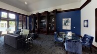 This single-floor clubhouse and leasing facility for Bristol Station presents a polished, upscale aesthetic defined by a cohesive navy blue and dark mahogany color palette throughout. The entry area features gleaming dark tile flooring, white structural columns with rich wood baseboards, and a welcoming "Welcome Home" mat, setting an elegant tone from the moment of arrival. A popcorn machine near the entrance adds a hospitable, community-focused touch.

The main lounge area commands attention with soaring vaulted ceilings, recessed lighting, and plush gray sofas accented with navy and geometric-patterned throw pillows. Tall windows dressed in wood blinds and navy draping flood the space with natural light while offering views of an outdoor pool area. A floor-to-ceiling dark mahogany built-in entertainment wall, complete with open display shelving, crown molding detail, and a centrally mounted television, serves as a dramatic focal point. Adjacent seating includes navy velvet chairs arranged around a glass-top table, reinforcing the sophisticated tone.

A dedicated business center offers a tray ceiling, dual computer workstations positioned beneath natural light-filled windows, black seating, and curated wall art, providing a functional yet design-forward workspace. The patterned charcoal carpet runs consistently throughout, unifying the various areas into a cohesive and refined overall environment.
