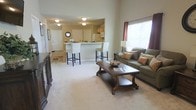 This single-story residence showcases an open-concept layout that integrates the living and kitchen areas into one cohesive, flowing space. The living room is appointed with plush wall-to-wall carpeting in a soft neutral tone, providing a warm and comfortable foundation. A generously sized green sofa adorned with decorative pillows anchors the seating area, complemented by an ornate dark wood coffee table that adds a traditional, elegant touch. A media console positioned along one wall offers additional storage and display space. Natural light filters in through a window dressed with rich burgundy curtains, brightening the room.

The kitchen is open to the living area and features a raised breakfast bar with white upholstered bar stools, making it an ideal casual dining or entertaining spot. Light wood cabinetry lines the kitchen walls, paired with white appliances and ample counter space. A circular mirror adds a decorative accent near the entry. The ceiling is fitted with flush-mount light fixtures that provide consistent, even illumination throughout. The neutral wall color and cohesive design elements create a clean, welcoming ambiance across the entire space.