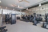 Located at 13900 Panay Way in Marina del Rey, California, this fitness center offers a spacious and thoughtfully designed workout environment spread across a single open floor. The room is bathed in natural light streaming through large, floor-to-ceiling glass windows and doors along one wall, providing views of the surrounding outdoor courtyard and palm tree-lined surroundings — a hallmark of the Southern California lifestyle.

The interior features a clean, modern aesthetic with neutral-toned carpeting, crisp white walls, and exposed industrial-style ductwork overhead that adds an urban edge to the otherwise polished space. Ceiling fans and pendant lighting contribute to both comfort and ambiance throughout the room.

The fitness center is comprehensively equipped for a full range of workouts. A dedicated cardio zone houses multiple treadmills, elliptical trainers, stair climbers, and stationary bikes. A strength training area includes a multi-function cable machine, adjustable benches, and a tiered dumbbell rack with a full weight selection. Floor-to-ceiling mirrored walls along one side of the room visually expand the space while allowing users to monitor their form. A modern staircase with a sleek cable-rail system leads to an upper level, and a framed artwork piece adds a refined, curated touch to the bright, open walls.