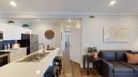 This single-floor residence in the US offers a well-appointed layout with modern finishes and a warm, cohesive aesthetic. The open-concept living and kitchen area serves as the heart of the home, where rich hardwood-style flooring, crown molding, and recessed lighting create an inviting atmosphere. The kitchen is equipped with stainless steel appliances, crisp white shaker cabinets, a striking navy blue tile backsplash, and a quartz countertop peninsula with bar seating — ideal for casual dining or entertaining. The living area provides comfortable space for relaxation, with a private exterior door that allows for natural light and outdoor access. The bedrooms offer a calm retreat with soft neutral tones, generous floor space, and ample closet access. The primary bathroom features white shaker-style cabinetry, a large vanity with quartz countertops, a tall linen tower, and a full-length decorative mirror, lending an open and airy feel. A second full bathroom echoes the clean, white palette with a tub-shower combination and stylish wood-framed wall accents. Throughout the home, consistent hardwood-style flooring, neutral gray walls, and white trim tie each space together with a polished, contemporary look.