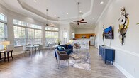 Located at Hawthorne Groves in Orlando, Florida, this single-floor community gathering space offers a well-appointed and versatile open-plan layout designed for comfort and functionality. The interior is anchored by light wood-tone flooring that flows seamlessly throughout, complemented by crisp white wainscoting, crown molding, and neutral wall tones that lend a clean, polished aesthetic.

The lounge area features a tray ceiling with dual ceiling fans, recessed lighting, and an elegant sofa and seating arrangement centered on an abstract area rug. A wall-mounted TV is flanked by decorative metalwork sconces above a bold navy console cabinet, adding a contemporary accent to the space.

Generous windows and transom windows line multiple walls, flooding the interior with natural light and offering views of the surrounding greenery and outdoor amenities. French doors provide a seamless transition to an adjacent foyer area, which showcases additional windows and refined trim details.

A dedicated dining area accommodates multiple tables and chairs, positioned to take full advantage of the panoramic window views. The fully equipped kitchen features warm wood cabinetry, light quartz-style countertops, stainless steel appliances including a refrigerator, dishwasher, and microwave, making it well-suited for community events and gatherings.