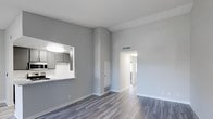 This single-floor space showcases a clean, modern design with a neutral grey and white color palette that creates a bright, airy atmosphere. The living area is distinguished by vaulted ceilings that add a sense of height and openness to the space. Durable wood-look plank flooring in a warm grey tone runs continuously throughout, providing a cohesive and contemporary aesthetic.

A pass-through window connects the kitchen to the living area, offering convenient sight lines between the two spaces. The kitchen features grey cabinetry, light countertops, and under-cabinet lighting, giving it a sleek, functional appearance. A base-height countertop ledge at the pass-through serves as an informal bar or serving area.

A central hallway leads to additional rooms, with an arched entryway adding a subtle architectural detail. The walls are finished in a smooth grey tone with crisp white baseboards and trim. Ceiling vents and floor-level vents indicate a central HVAC system throughout. Overall, the space presents a well-maintained, move-in-ready interior with a minimalist and modern character.