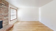 This single-floor apartment in Beaverton, Oregon offers a well-organized layout accommodating three bedrooms and two full bathrooms. The living room serves as a central gathering space, highlighted by a striking floor-to-ceiling brick fireplace flanked by two large windows with wood-trimmed frames, allowing natural light to pour across the light wood laminate flooring. The open-concept kitchen flows directly from the living area and features warm wood cabinetry, a white appliance suite including a refrigerator and electric range, a built-in peninsula with a white countertop ideal for casual dining or food preparation, and a ceiling fan overhead. A window above the sink provides additional natural light.

The three bedrooms are carpeted and each include windows overlooking greenery, baseboard heaters, and textured ceilings. The spacious primary bedroom offers ample room for furnishings. The two bathrooms are clean and functional—one features a full tub and shower combination with white tile surround, while the other includes a standalone walk-in shower unit with built-in shelving. Throughout, neutral wall tones, wood-trimmed accents, and consistent flooring materials create a cohesive and practical living environment.
