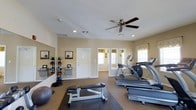 This fitness room is designed to support a well-rounded exercise routine within a compact, functional layout. The space is finished with neutral beige walls and carpeted flooring, creating a clean and approachable atmosphere. A ceiling fan mounted at the center of the white ceiling provides air circulation throughout workouts, complementing the ventilation system supported by visible ceiling vents.

The room is equipped with SportsArt cardio machines, including a treadmill and an elliptical trainer, positioned along one side of the space. A weight bench with a barbell setup occupies the central floor area, while a stability ball and additional free weights are available nearby. Along one wall, a vertical rack holds an organized assortment of medicine balls in varying sizes, accompanied by framed informational posters. A large mirror is mounted beside the rack, visually expanding the space and allowing users to monitor their form.

Natural light filters in through multiple windows dressed with simple valances, brightening the room. A doorway connects to an adjacent windowed area, adding depth to the layout and enhancing the overall sense of openness.
