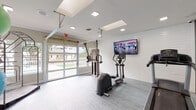 This fitness center is a thoughtfully designed, single-floor facility that offers a comprehensive workout environment. The space features light gray vinyl flooring throughout, white shiplap accent walls, and a bright ceiling equipped with recessed lighting and skylights that keep the interior well-lit and energizing.

The main workout area houses a variety of cardio equipment, including treadmills, an elliptical trainer, and a rowing machine, all positioned to face a large wall-mounted flat-screen television. A multi-functional cable resistance machine provides options for strength training, rounding out the cardiovascular offerings.

A dedicated studio area is enclosed with floor-to-ceiling glass partitions, creating a separate, open space ideal for stretching, yoga, or group fitness activities. This area is further equipped with a rack of medicine balls and stability balls in various sizes, offering versatile functional training options.

One of the most distinctive architectural features of the fitness center is the large, glass-panel garage-style doors that open to reveal views of the surrounding outdoor amenity areas, including a pool and landscaped grounds. This design allows for abundant natural light and fresh air circulation, creating an inviting and open atmosphere. The overall aesthetic is clean, modern, and conducive to an effective workout experience.