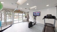This fitness center is a thoughtfully designed, single-floor facility that offers a comprehensive workout environment. The space features light gray vinyl flooring throughout, white shiplap accent walls, and a bright ceiling equipped with recessed lighting and skylights that keep the interior well-lit and energizing.

The main workout area houses a variety of cardio equipment, including treadmills, an elliptical trainer, and a rowing machine, all positioned to face a large wall-mounted flat-screen television. A multi-functional cable resistance machine provides options for strength training, rounding out the cardiovascular offerings.

A dedicated studio area is enclosed with floor-to-ceiling glass partitions, creating a separate, open space ideal for stretching, yoga, or group fitness activities. This area is further equipped with a rack of medicine balls and stability balls in various sizes, offering versatile functional training options.

One of the most distinctive architectural features of the fitness center is the large, glass-panel garage-style doors that open to reveal views of the surrounding outdoor amenity areas, including a pool and landscaped grounds. This design allows for abundant natural light and fresh air circulation, creating an inviting and open atmosphere. The overall aesthetic is clean, modern, and conducive to an effective workout experience.