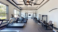 Located at 10095 Washington Blvd N in Laurel, Maryland, this fitness center offers a thoughtfully designed, single-floor exercise space that combines functionality with modern aesthetics. The room is characterized by expansive floor-to-ceiling windows along multiple walls, flooding the interior with natural light and offering open views of the surrounding greenery and streetscape.

The space features a refined architectural design, highlighted by a coffered ceiling with recessed lighting and a ceiling fan, dark crown molding trim, and clean white and light gray walls that create a bright, energizing atmosphere. Durable speckled rubber flooring in a dark blue-gray hue covers the entire area, providing both safety and a polished, professional appearance.

The exercise room is comprehensively equipped for a full-body workout. Cardio equipment includes multiple treadmills and an elliptical trainer, neatly arranged along the windowed wall. Strength training is well-represented with a cable pulley machine, a leg extension/curl station, a lat pulldown machine, a Smith machine, free weights, and adjustable benches. A wall-mounted flat-screen television adds a convenient entertainment option during workouts.

Glass double doors connect the fitness area to an adjacent interior space, enhancing the open, accessible feel of the facility. The overall ambiance is clean, modern, and motivating.