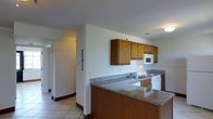 This single-floor space presents a practical and straightforward layout centered around a well-equipped kitchen. The kitchen serves as a focal point, featuring a peninsula with granite-style countertops that extend along the perimeter walls, offering ample prep and workspace. Warm-toned wood upper cabinetry provides generous storage, while a white electric range, built-in microwave, and stainless steel sink contribute to the kitchen's functional design. Light tile flooring runs throughout the space, lending a clean, cohesive look that is both easy to maintain and visually consistent. The white walls and standard ceiling height create a bright, neutral backdrop throughout. A hallway visible from the kitchen leads to additional areas of the space, suggesting a connected, flowing floor plan. The overall ambiance is clean and utilitarian, designed with everyday functionality in mind. The kitchen peninsula also doubles as a casual dining or gathering area, adding versatility to the layout.