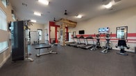 This fitness center offers a comprehensive workout environment designed to support a wide range of exercise routines. The main area is spacious and open, featuring durable rubber flooring throughout and a bold design aesthetic highlighted by red accent stripes along the walls and a striking reclaimed wood column detail that adds a warm, industrial character to the space. Ceiling fans and recessed lighting ensure the area remains comfortable and well-lit during workouts.

The cardio zone is equipped with multiple treadmills, elliptical machines, and rowing machines, all positioned along the main wall and accompanied by wall-mounted televisions for entertainment. A dedicated strength training section includes a multi-functional Smith machine with an adjustable bench, a cable machine from Spirit Fitness, and a tiered dumbbell rack stocked with a full range of free weights. Additional benches and functional training equipment are spread throughout the open floor area.

Natural light flows into the space through multiple windows fitted with plantation shutters, offering views of the surrounding exterior. The separate strength room benefits from windows on multiple walls, creating a bright, airy feel. The overall ambiance is modern and motivating, combining practical equipment placement with thoughtful design details.