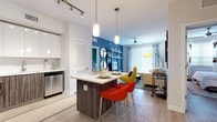 This single-floor residence showcases a contemporary design with a cohesive open-concept layout connecting the kitchen, dining, and living areas. The kitchen is a standout feature, equipped with a full suite of stainless steel appliances including a French door refrigerator, range, microwave, and dishwasher. A two-toned kitchen design pairs dark wood-grain lower cabinetry with glossy white upper cabinets, complemented by white countertops and light wood-style flooring that extends throughout the unit. A central island with seating serves as both a dining and gathering space, illuminated by pendant lighting and adjustable track lighting overhead.

The living area features a bold blue accent wall with built-in shelving, a ceiling fan, and large windows that invite natural light. The bedroom continues the vibrant accent wall theme with a striking green feature wall, ceiling fan, and a window providing additional natural light. A second bedroom presents a clean, neutral palette. The bathroom includes a soaking tub with full tile surround in a neutral gray tone. Throughout the space, light wood-style flooring, modern fixtures, and thoughtful lighting combine to create a bright, stylish, and well-appointed interior.