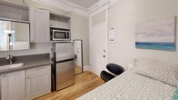 Located at 1010 10th St in Sacramento, CA, this compact studio unit is thoughtfully designed to maximize every square foot of its single-room footprint. The space is organized into distinct functional zones, making efficient use of its cozy layout. Warm-toned hardwood flooring runs throughout, lending a clean and cohesive feel to the interior. The sleeping area accommodates a twin bed with under-bed storage, complemented by a freestanding white wardrobe unit with open shelving — providing practical storage without overwhelming the space. Coastal-themed wall art adds a light, calming aesthetic to the soft sage-green walls. The kitchenette area is well-appointed with light gray shaker-style cabinetry, open shelving, a stainless steel sink with countertop space, a full-size stainless steel refrigerator, and a microwave — offering essential cooking amenities in a compact footprint. A full-length mirror and a sleek black accent chair round out the furnishings. Natural light enters through multiple windows fitted with blinds, brightening the space considerably. Crown molding details add a subtle architectural character. A wall-mounted climate control unit ensures year-round comfort. This efficiently designed space is well-suited for a single occupant prioritizing location and functionality in the heart of Sacramento.