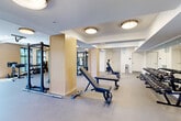 Located at 110 E 36th Street in Charlotte, NC, this well-appointed fitness center offers a generously sized, single-floor exercise facility designed to accommodate a comprehensive range of workout routines. The space is thoughtfully divided into distinct functional zones, creating an organized and efficient environment for all fitness levels.

The strength training area is equipped with power racks, adjustable benches, and an extensive dumbbell rack, all set against large mirrored walls that enhance the sense of space and allow users to monitor their form. Light wood-clad columns provide architectural interest and help define the zones within the open layout.

The cardio zone features multiple Life Fitness treadmills, stair climbers, stationary bikes, air bikes, and rowing machines, all positioned to take advantage of large, multi-pane windows that flood the area with natural light and offer views of the surrounding neighborhood. A glass garage-style door on one end opens the space further, connecting it visually to the outdoors.

Ceiling fans are strategically placed throughout, complementing the flush-mount lighting to maintain a comfortable environment. The durable rubber-style flooring provides both safety and functionality across the entire space. The overall aesthetic is clean and modern, with a neutral white and warm wood palette that creates a bright, motivating atmosphere conducive to focused training.