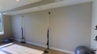 This single-floor fitness space is designed to support functional training with a clean, minimalist aesthetic. The room features smooth, neutral gray walls that create a calm and focused environment, complemented by warm wood-look flooring that adds a welcoming yet practical surface underfoot. The ceiling is finished with a flat, light-toned surface and includes a flush panel fixture that provides even ambient lighting throughout the space.

A key feature of this room is the ceiling-mounted anchor hardware, from which TRX-style suspension training straps hang, enabling a wide range of bodyweight exercises. Multiple suspension systems are visible, suggesting the space can accommodate several users simultaneously. The anchor points are securely fixed, indicating the room has been purpose-built or thoughtfully retrofitted for fitness use.

White baseboards frame the perimeter of the room, contributing to the tidy, well-maintained appearance. The open floor plan allows for free movement, making the space adaptable for stretching, strength training, and suspension-based workouts. Overall, the space presents a functional and organized training environment suited for individual or small group use.