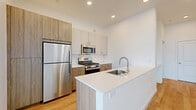 This single-floor unit presents a clean, contemporary design throughout, characterized by crisp white walls, light hardwood flooring, and recessed lighting that create a bright and cohesive aesthetic. The open-concept kitchen serves as a central feature, equipped with a two-toned cabinetry system combining white upper cabinets and wood-finish lower cabinets, a white subway tile backsplash, and a full suite of stainless steel appliances including a gas range, over-the-range microwave, and a full-size refrigerator with matching tall pantry cabinet. A white quartz kitchen island provides additional prep space and includes an undermount sink with a modern arc faucet. The living area is generously proportioned, featuring large windows and a glass balcony door that allow abundant natural light and outdoor views. Bedroom spaces are well-sized with sliding-door closets offering built-in hanging storage. The bathroom is finished with a glass-enclosed walk-in shower, chrome fixtures, and wood-look tile flooring, maintaining the unit's consistent modern style. The overall layout is functional and well-organized.