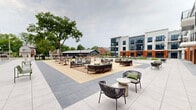 Located at 1222 South Ave in Plainfield, NJ, this contemporary outdoor courtyard space is set within a multi-story building complex that combines brick and panel facade elements for a modern, urban aesthetic. The courtyard serves as a central amenity hub, thoughtfully designed to encourage outdoor activity and social interaction.

The space features multiple lush green lawn areas maintained with vibrant artificial turf, providing room for recreational activities such as cornhole. Wide-format porcelain tile paving connects the various zones seamlessly, creating a clean and polished hardscape throughout.

Seating is distributed across the courtyard in various configurations, including sleek green metal benches and modular lounge furniture arranged around gathering areas. Umbrella-shaded bistro tables offer additional spots for relaxation. The ground-floor perimeter of the building is lined with floor-to-ceiling glass storefronts and sliding doors, allowing interior spaces to open directly onto the courtyard.

Upper-level balconies with dark metal railings overlook the courtyard, reinforcing the sense of community and connectivity between indoor and outdoor environments. A raised perimeter terrace along one edge provides views of the surrounding Plainfield neighborhood, framed by brick knee walls and metal railings. The overall ambiance is open, airy, and well-appointed, making this courtyard a standout communal feature of the property.