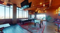 This dynamic, elevated game room delivers an expansive entertainment experience with a bold, eclectic design aesthetic. The space is divided into distinct activity zones, each thoughtfully appointed for both active play and social lounging. Floor-to-ceiling windows line the perimeter, flooding the room with natural light and offering stunning panoramic views of the city skyline that stretch to the horizon.

The main gaming area features an air hockey table, foosball table, and a billiards table, all anchored by richly patterned area rugs atop wide-plank hardwood flooring. Overhead, exposed ductwork and industrial-style pendant lighting lend the space a lively, urban character, while a wall-mounted flat-screen TV adds a multimedia element.

An arcade zone showcases classic machines including dual Sonic All-Stars basketball shooters, a skee-ball lane, and retro cabinets, complemented by casual dining tables with upholstered stools. A long plush banquette in deep violet velvet lines one wall, accented by framed artwork, small cocktail tables, and warm sconce lighting against rich terracotta walls.

A separate lounge area features contemporary black leather seating, a patterned area rug, and a built-in shelf display bathed in green accent lighting. Intimate seating nooks with plush armchairs positioned directly at the windows provide the perfect vantage point for taking in the breathtaking cityscape below.